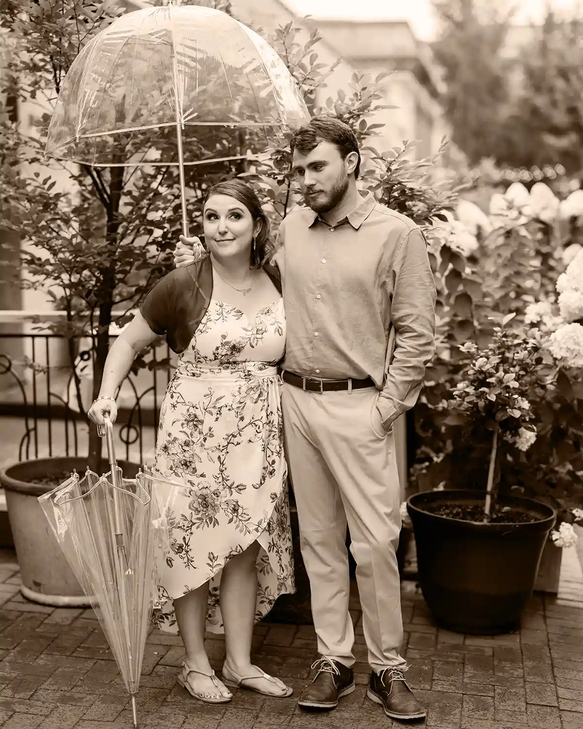 A young couple. The man is holding an umbrella and looks slightly annoyed. The woman is standing next to him with a sassy smile.